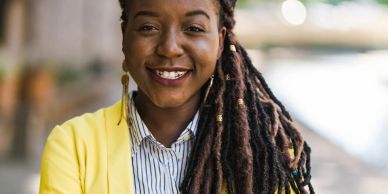 Smiling woman with long dreadlocks wearing a yellow blazer outdoors.