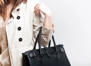 Woman in beige trench coat holding a black leather handbag.